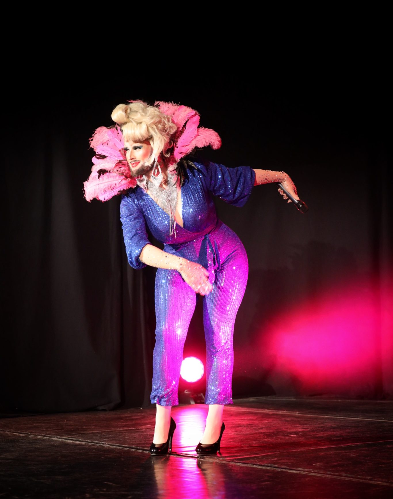 Person in glitzerndem blauen Outfit mit rosa Blumen, posiert dramatisch auf der Bühne.